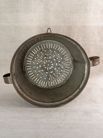 Antique French passoire in tinned sheet iron with rosette base and twin handles on neutral fabric.