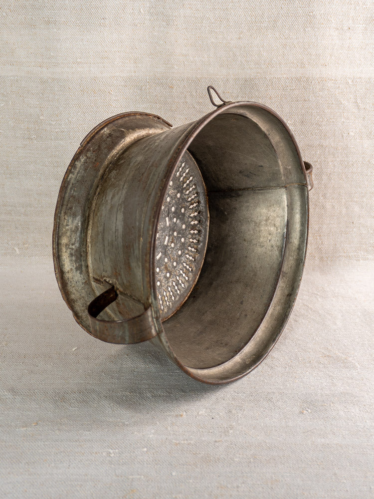 Antique French tinned sheet iron passoire with side handles and pierced rosette base on fabric background.