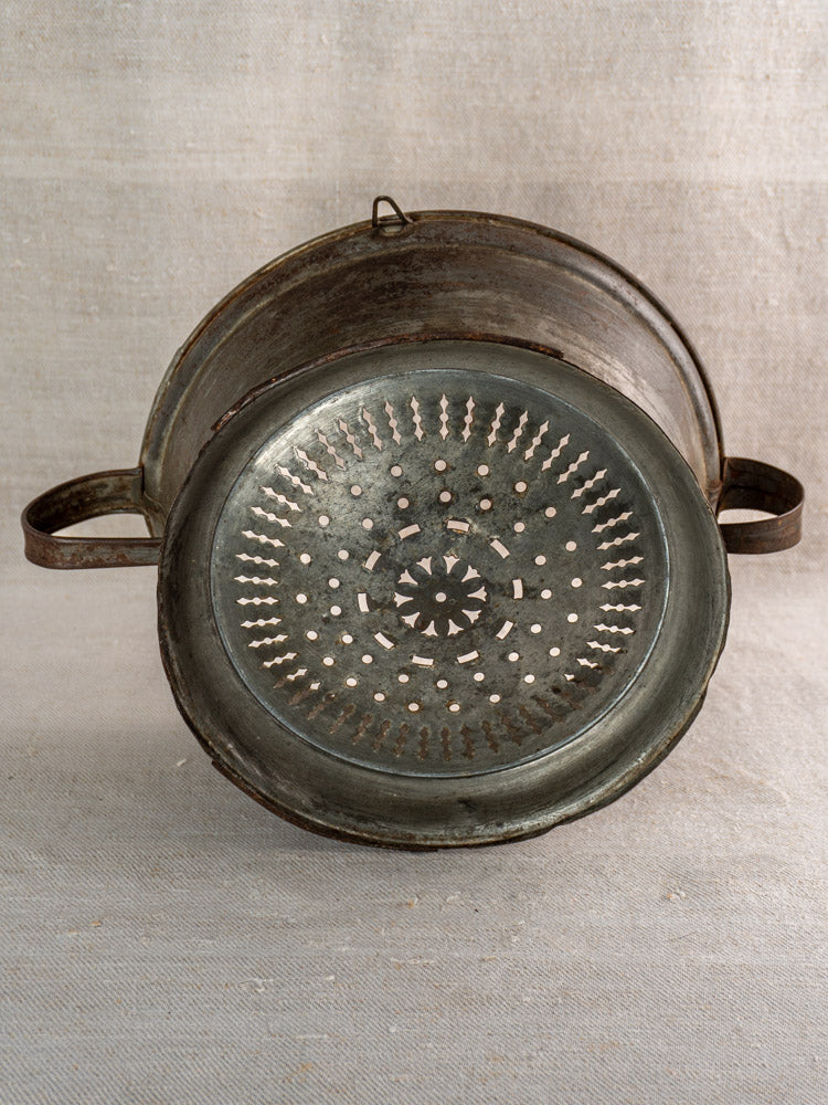 Antique French passoire in tinned sheet iron with twin handles, rosette base, on neutral fabric.