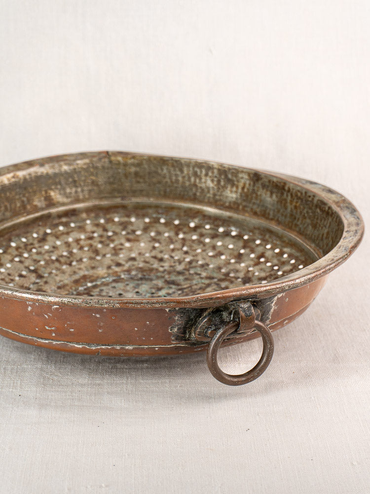 Antique French copper colander with hand-tinned interior and large side ring handle on light fabric background.