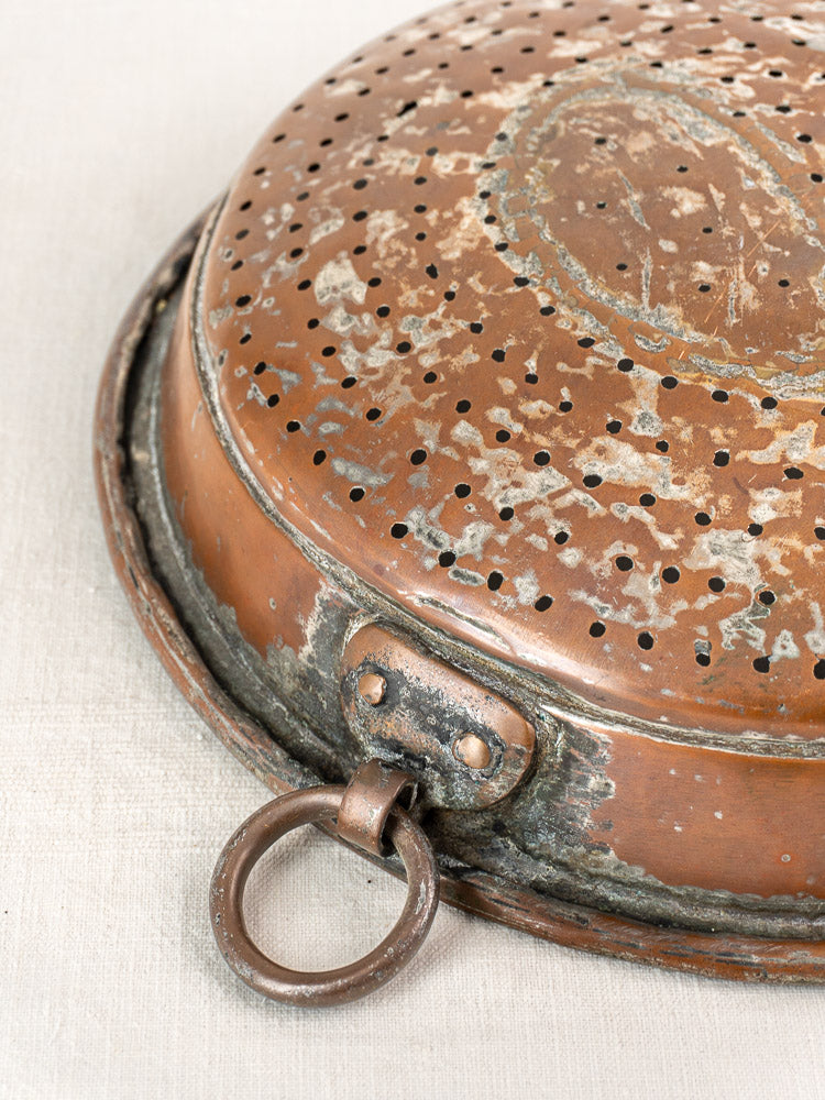Antique French copper colander with metal ring handle, tarnished patina, on a light fabric background.