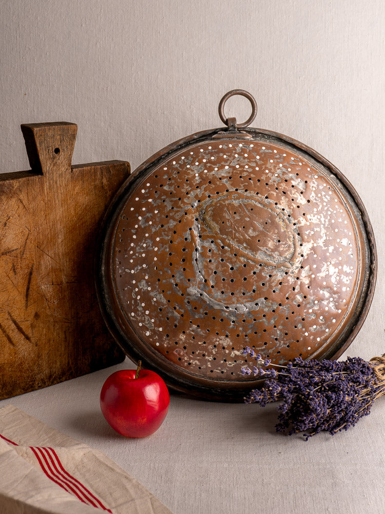 Antique French copper colander on light surface with wooden board, red apple, dried lavender, and striped cloth.