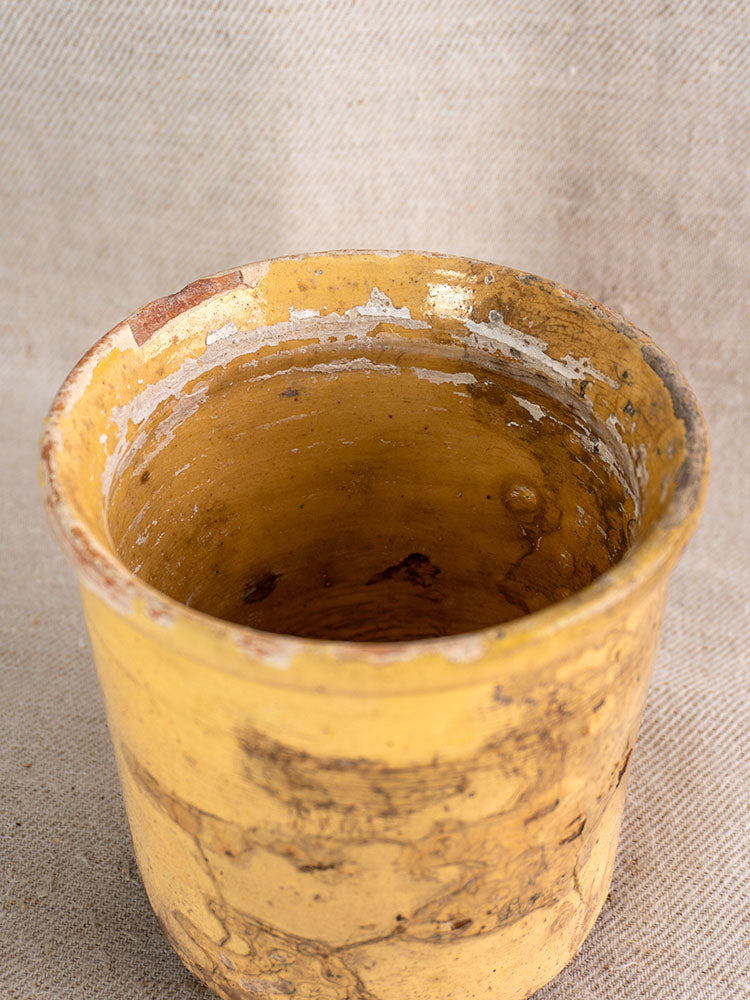 Antique French confit pot with weathered marbled yellow glaze and chipped rim on neutral fabric background.