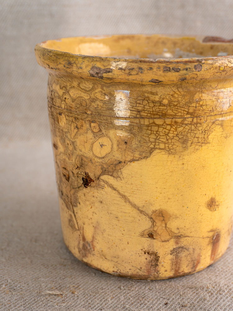Antique French confit pot with yellow glaze, cracked surface, and patina, set on neutral fabric background.
