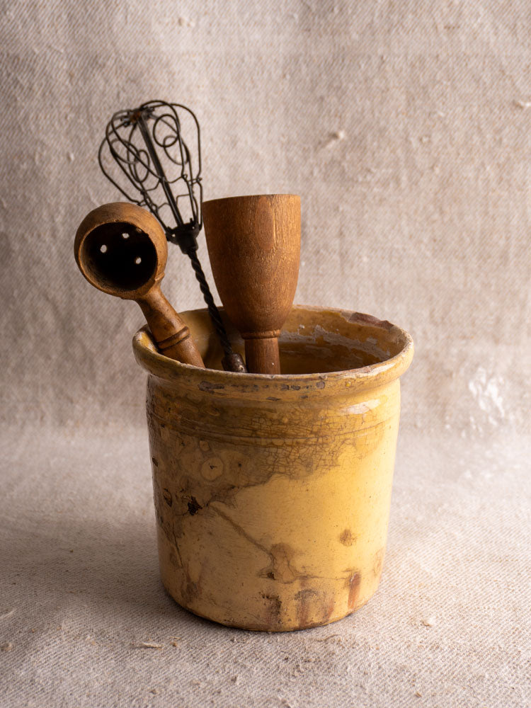 Antique French confit pot with yellow glaze holding wooden and metal utensils on beige textured fabric.