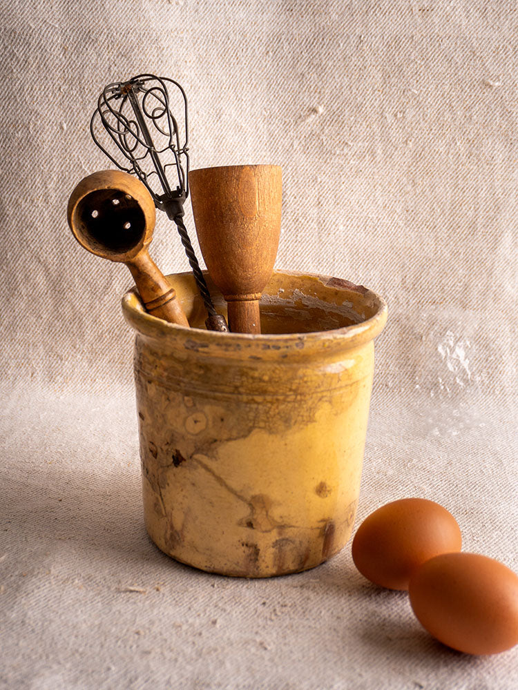 Antique French yellow-glazed confit pot with wooden utensils, displayed on textured fabric beside two brown eggs.