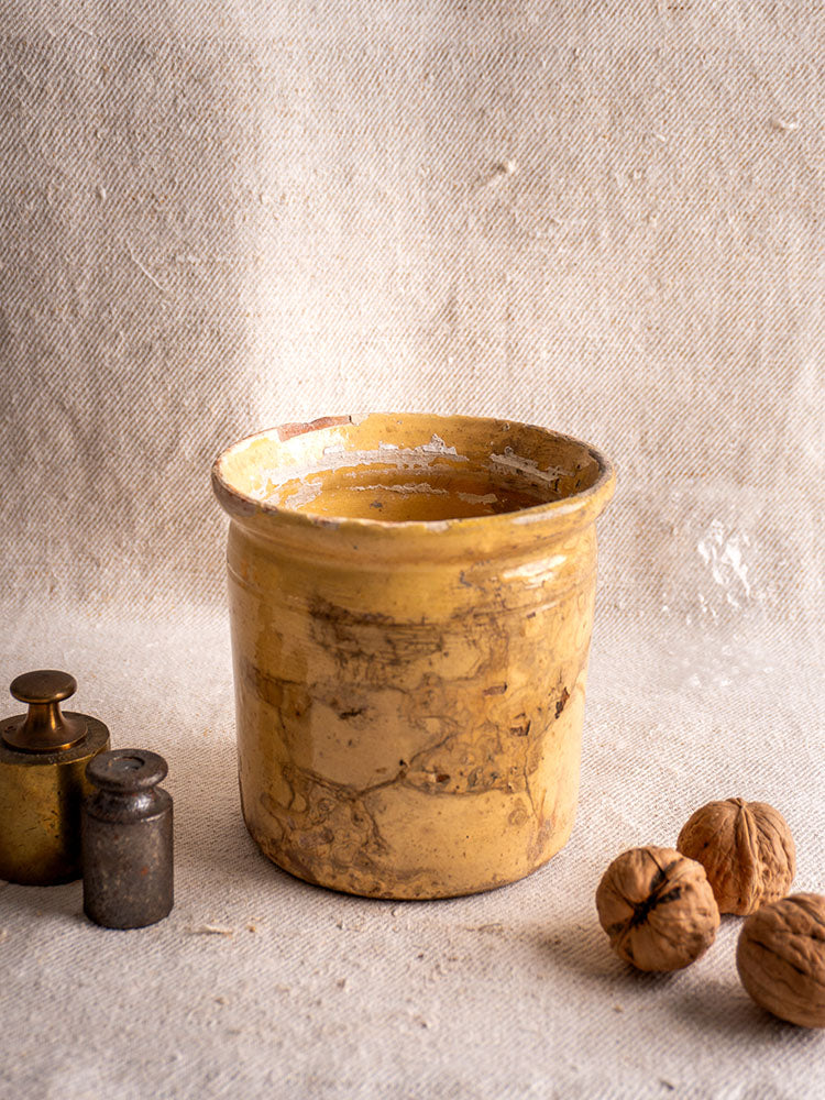 Antique French yellow glazed earthenware confit pot on beige cloth with metal weights and walnuts in soft natural light.