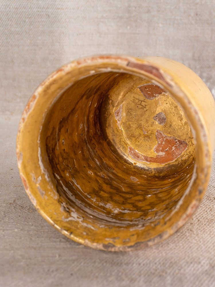 Antique French confit pot with aged yellow glaze, chips, and wear on a textured beige fabric background.