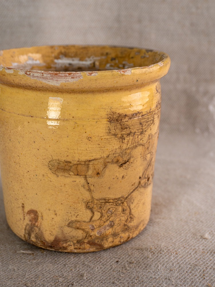 Antique French confit pot, 19th-century yellow glazed earthenware with rim chips, on textured beige fabric background.