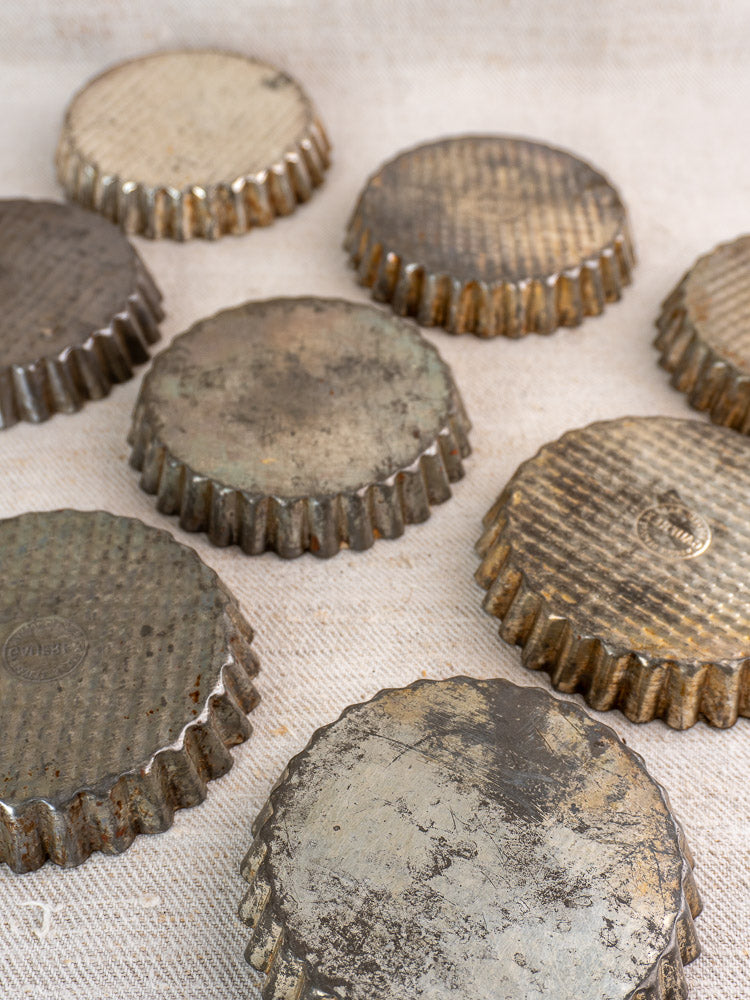 Nine vintage French tinned steel tart pans with worn fluted edges, patina, on a textured light fabric surface.