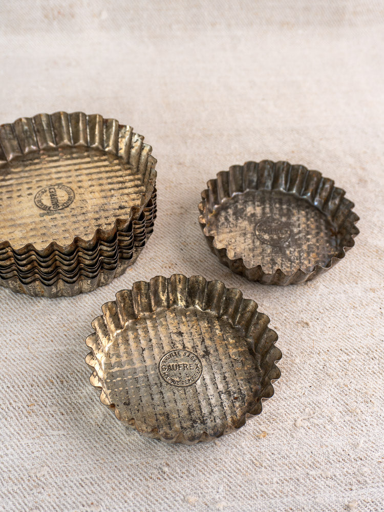 Stacked vintage tinned steel French baking molds with two tart pans, mid 20th century, on a light textured surface.