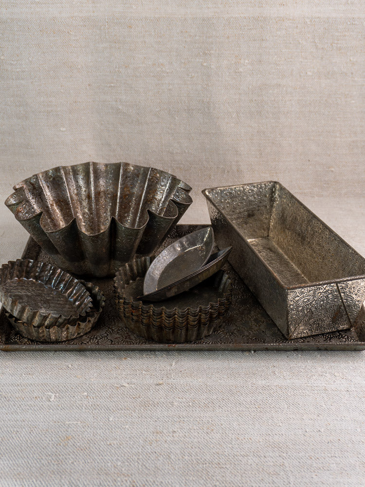 Antique tinned steel French tartlet molds, round and fluted, on ornate metal tray with neutral fabric backdrop.