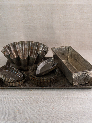 Antique tinned steel French tartlet molds, round and fluted, on ornate metal tray with neutral fabric backdrop.