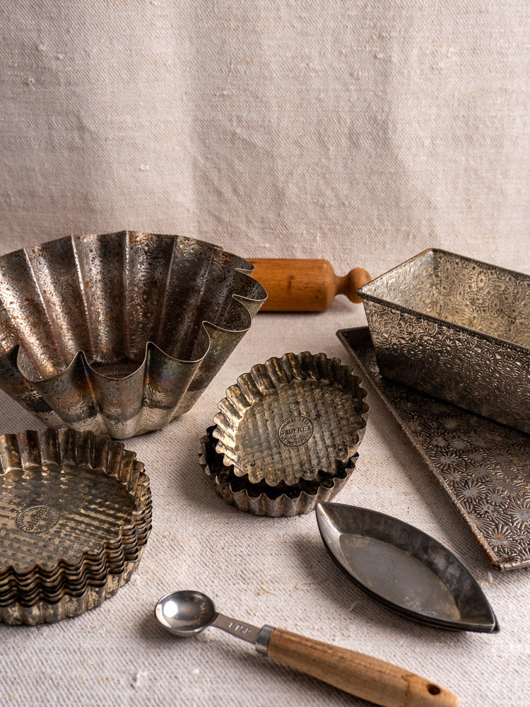 Mid-century tinned steel French bakery molds, rolling pin, loaf pan, and measuring spoon on beige fabric.