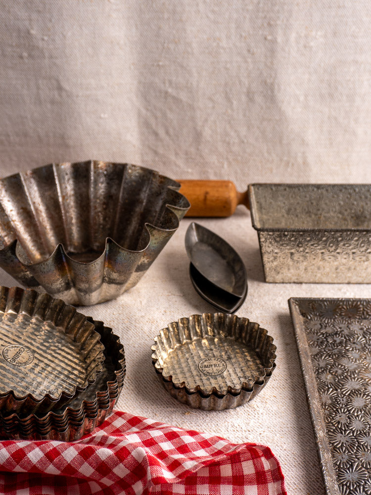 Mid-20th century tinned steel tart pans and loaf pan displayed on textured cloth with a red and white checkered towel.