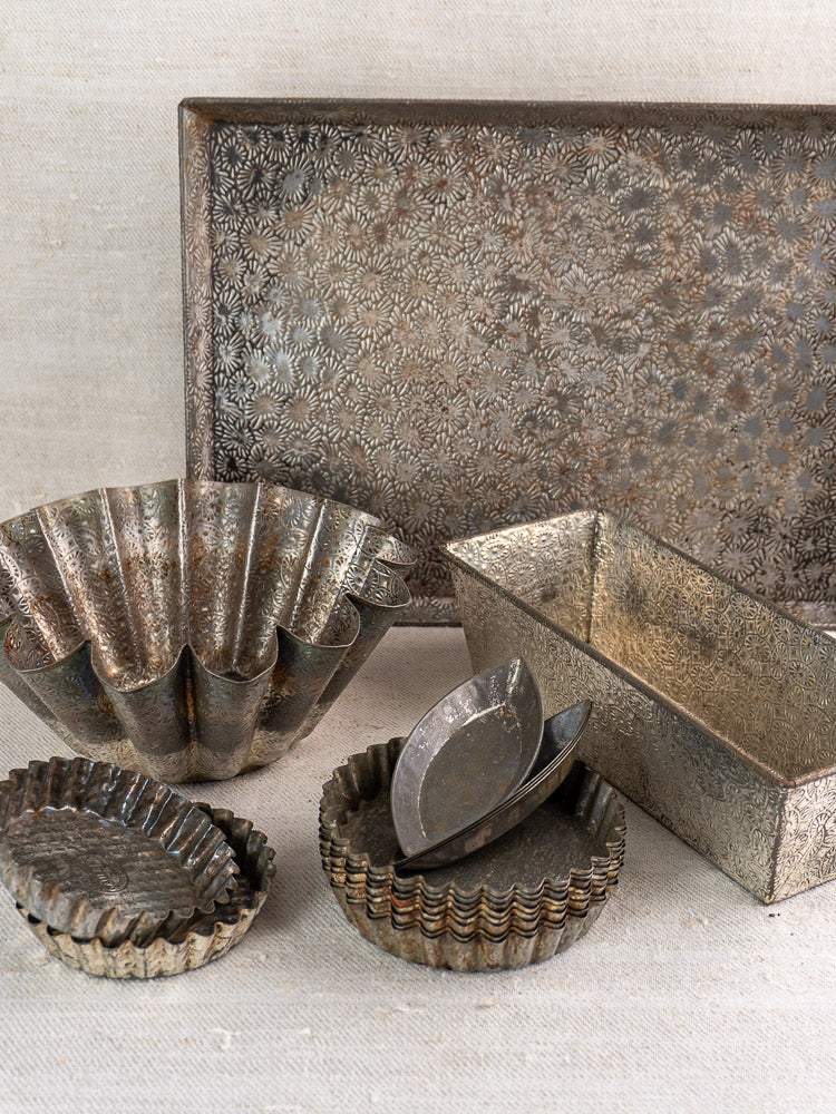 Mid-century French tinned steel tart pans, loaf pan, floral tray, and fluted mold on neutral fabric.
