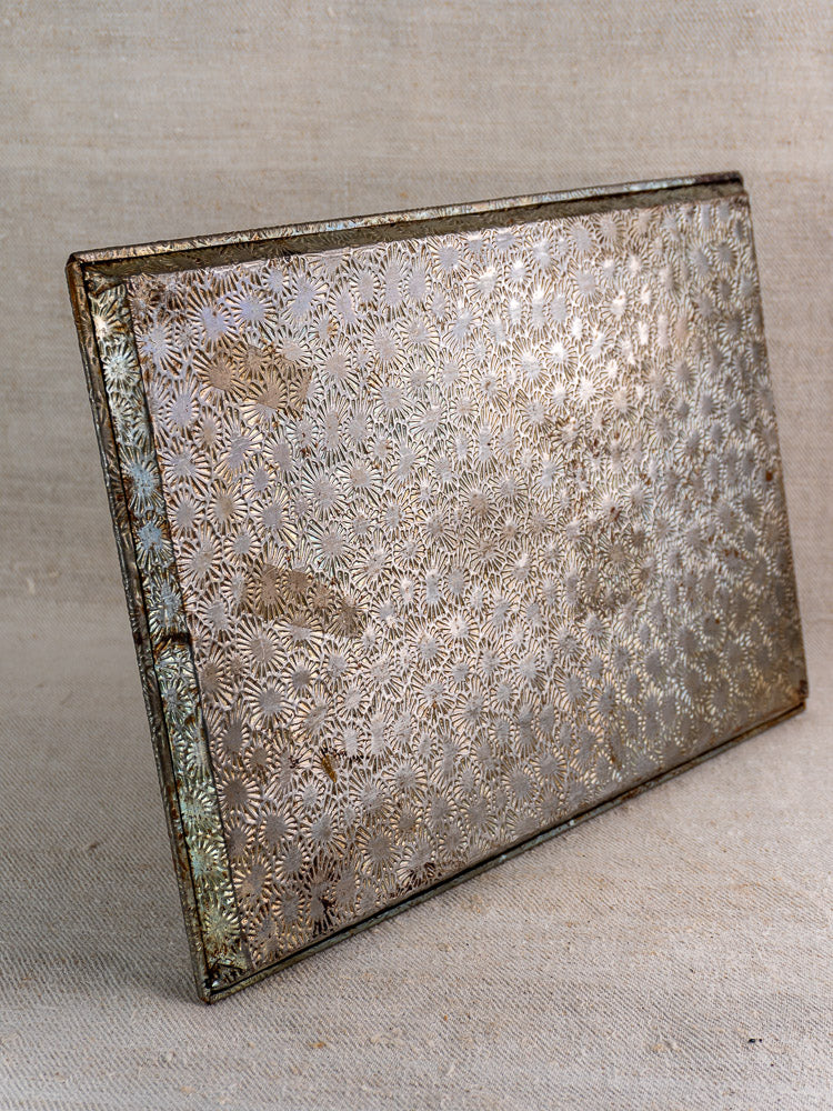 Mid-20th century French tinned steel box with embossed floral motif, slightly open on neutral fabric background.
