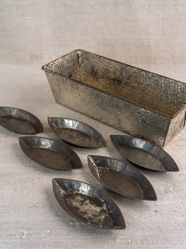Rectangular tinned steel loaf pan and six oval vintage French baking molds on textured beige fabric.