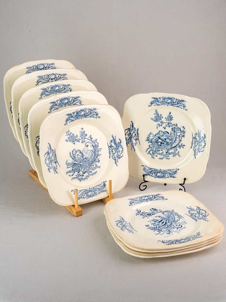 Set of 10 antique French Clairefontaine Louis XV plates with blue floral scrolls, in a wooden rack on light gray surface.