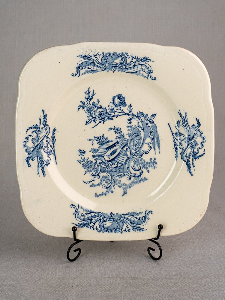 Square cream plate with blue Louis XV Terre de Fer floral design, on black stand, gray background.