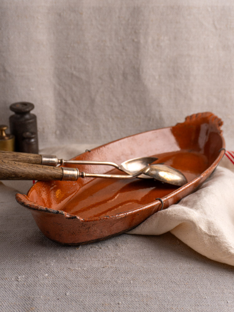 Late 19th-century French Saint-Uze oval terre vernissée baking dish with end handles on black stand, neutral fabric backdrop.