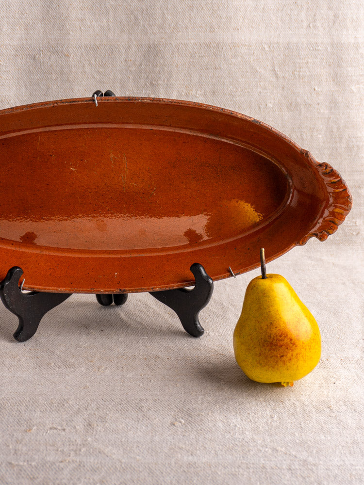 Antique French Saint-Uze Revol baking dish on black stand beside speckled yellow pear, against beige fabric background.