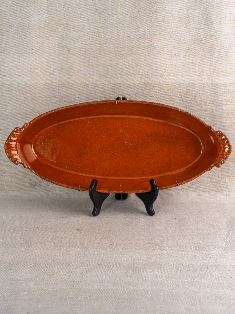 Late 19th-century French Saint-Uze oval terre vernissée baking dish with end handles on black stand, neutral fabric backdrop.