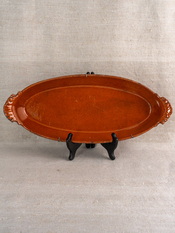 Late 19th-century French Saint-Uze oval terre vernissée baking dish with end handles on black stand, neutral fabric backdrop.