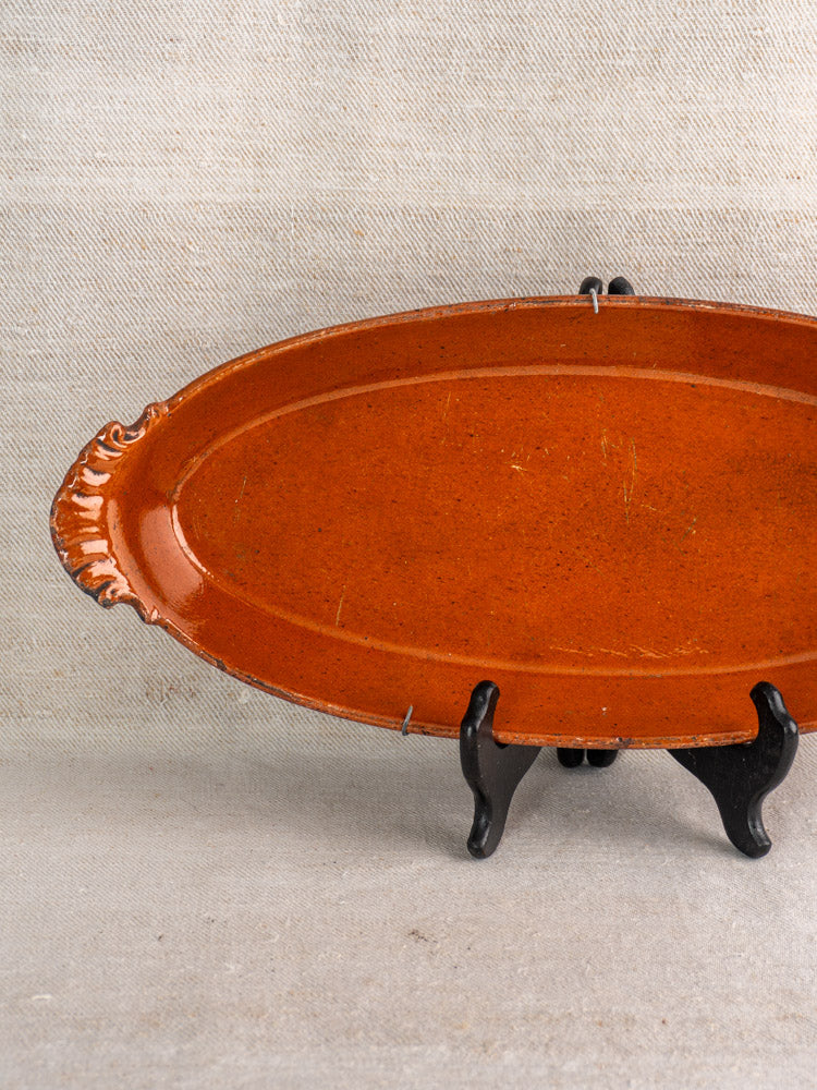 Antique French Saint-Uze Revol oval terre vernissée baking dish with ornate handle, on black stand, beige fabric backdrop.