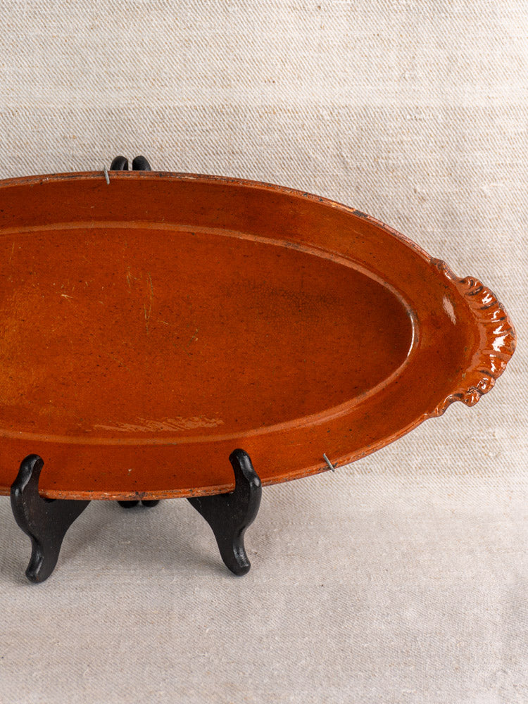 Antique oval Saint-Uze Revol terre vernissée baking dish with ornate handles, upright on black stand against neutral fabric.