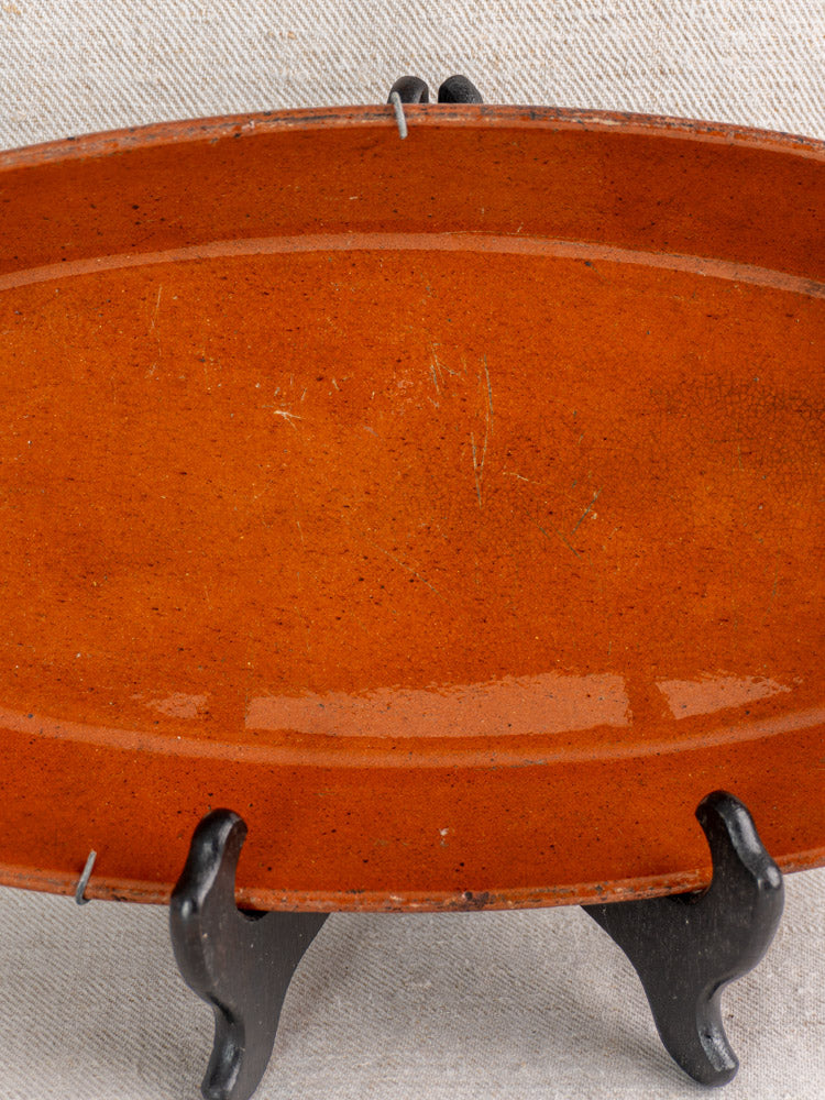 Antique Saint-Uze Revol French oval terre vernissée baking dish, reddish-brown glaze, on black stand with beige fabric backdrop.