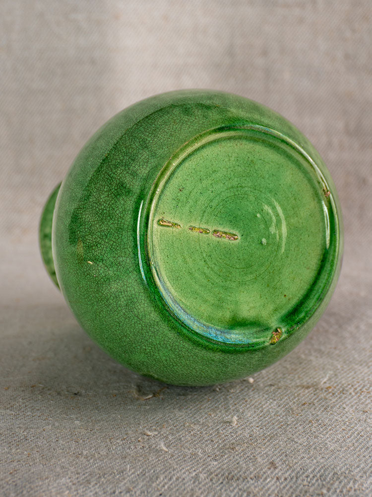 Antique French Dieulefit green glazed earthenware pitcher, glossy crackled base, on textured neutral fabric background.