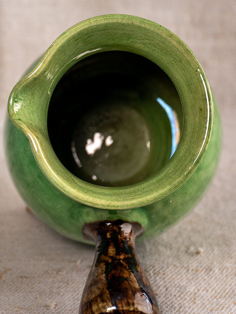 Antique French Dieulefit green glazed earthenware pitcher with dark marbled handle on textured light fabric.