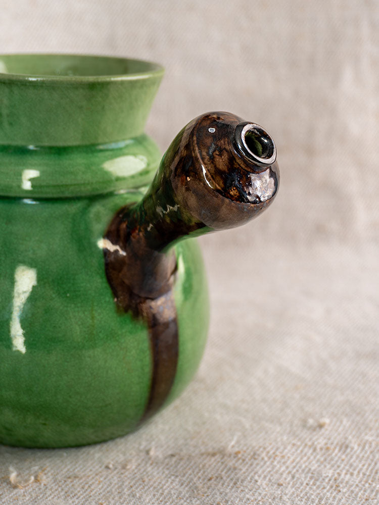 Antique French Dieulefit pitcher, late 19th c., green glazed earthenware with brown accents, on beige textured fabric.