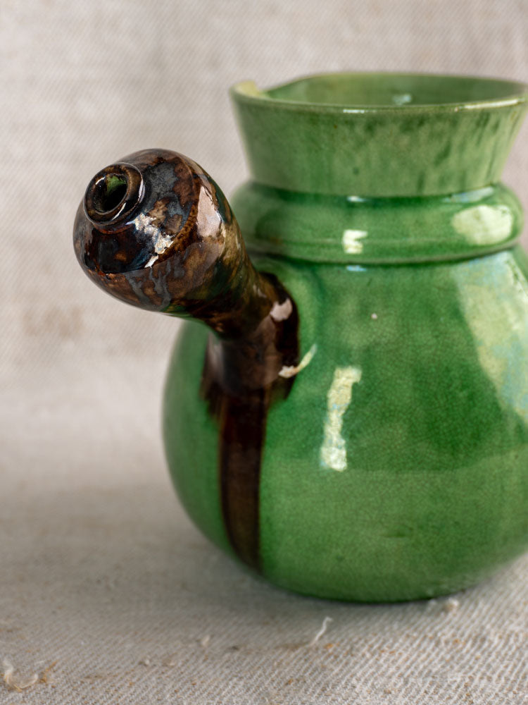 Antique French Dieulefit pitcher with green glazed earthenware body and brown spout on neutral fabric background.