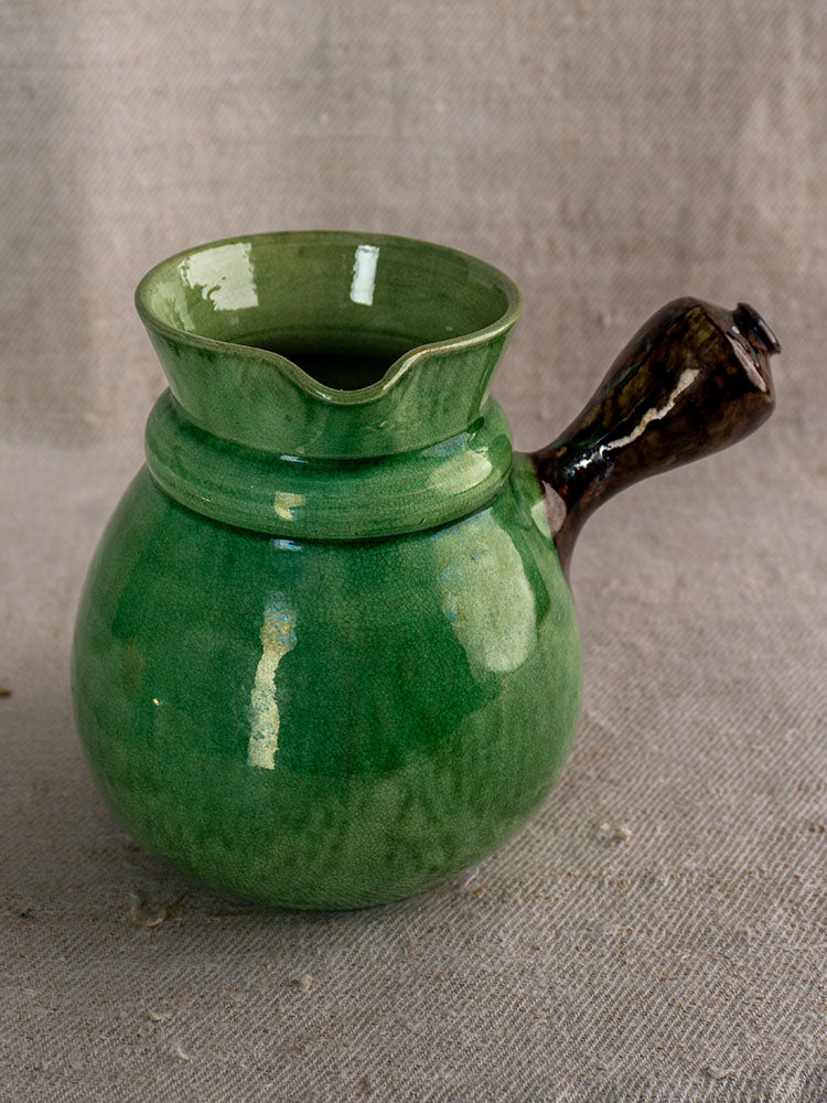 Antique French Dieulefit green glazed earthenware pitcher with brown handle on textured beige fabric.