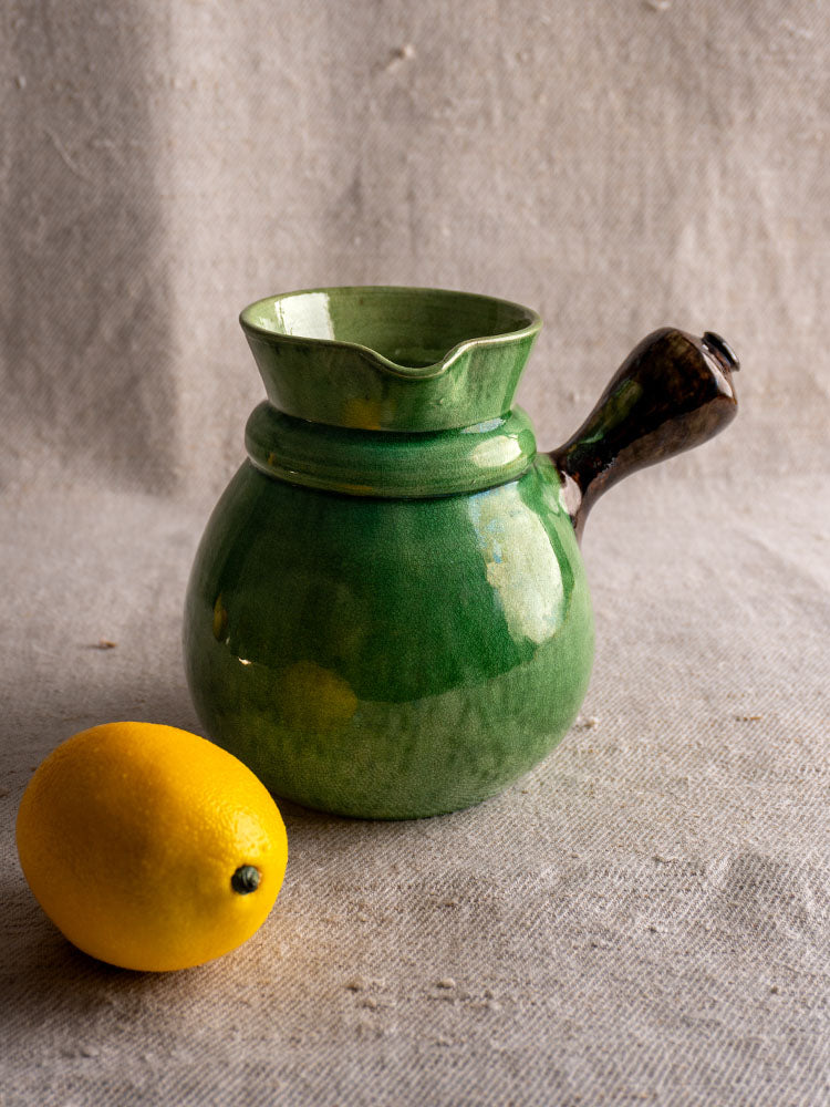 Antique French Dieulefit green glazed earthenware pitcher with dark handle beside a lemon on textured beige fabric.