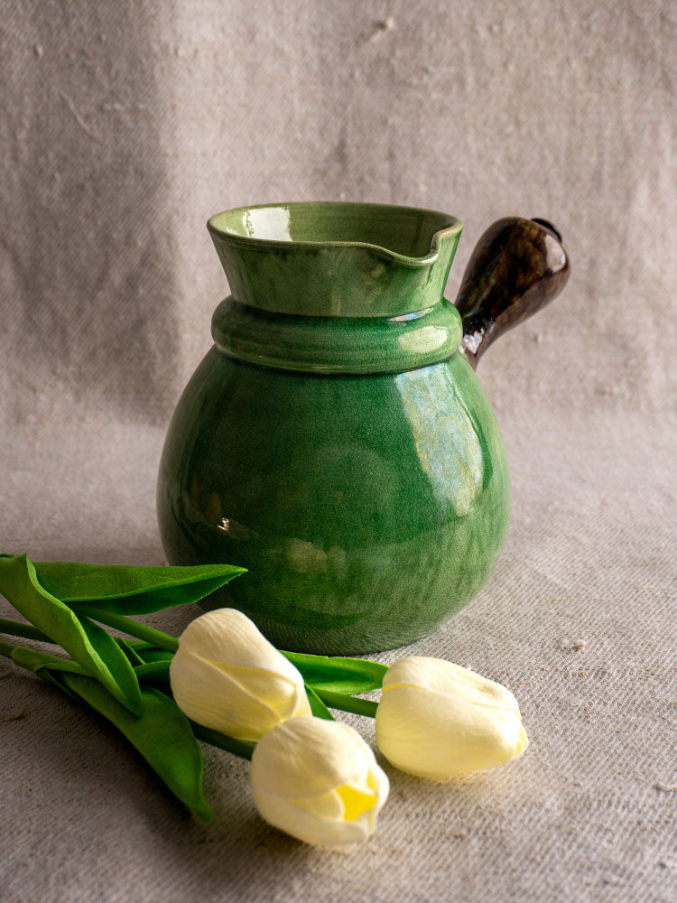 Antique French Dieulefit green glazed earthenware pitcher with dark handle on beige fabric, next to three white tulips.