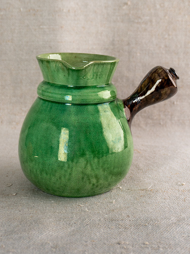 Antique French Dieulefit green glazed earthenware pitcher with wide spout and dark handle on beige fabric background.