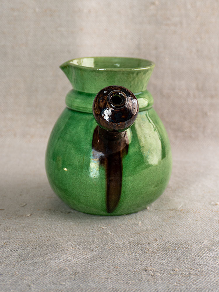 Antique French Dieulefit green glazed earthenware pitcher with dark brown spout on neutral textured fabric background.
