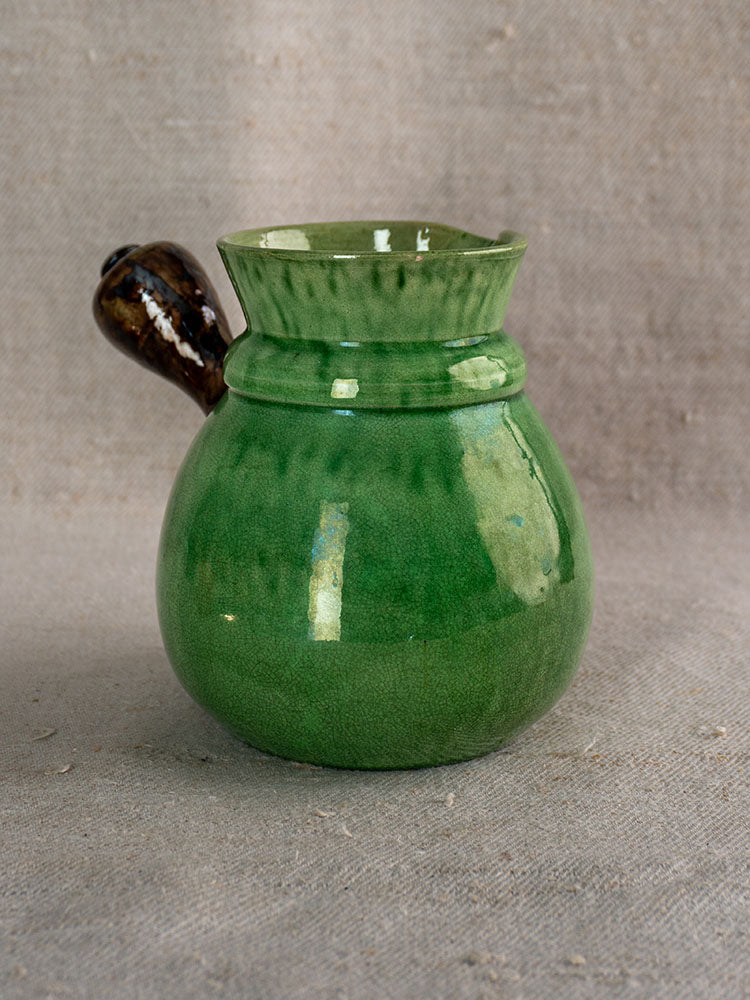 Antique French Dieulefit green glazed earthenware pitcher with round body and spout on beige textured fabric.