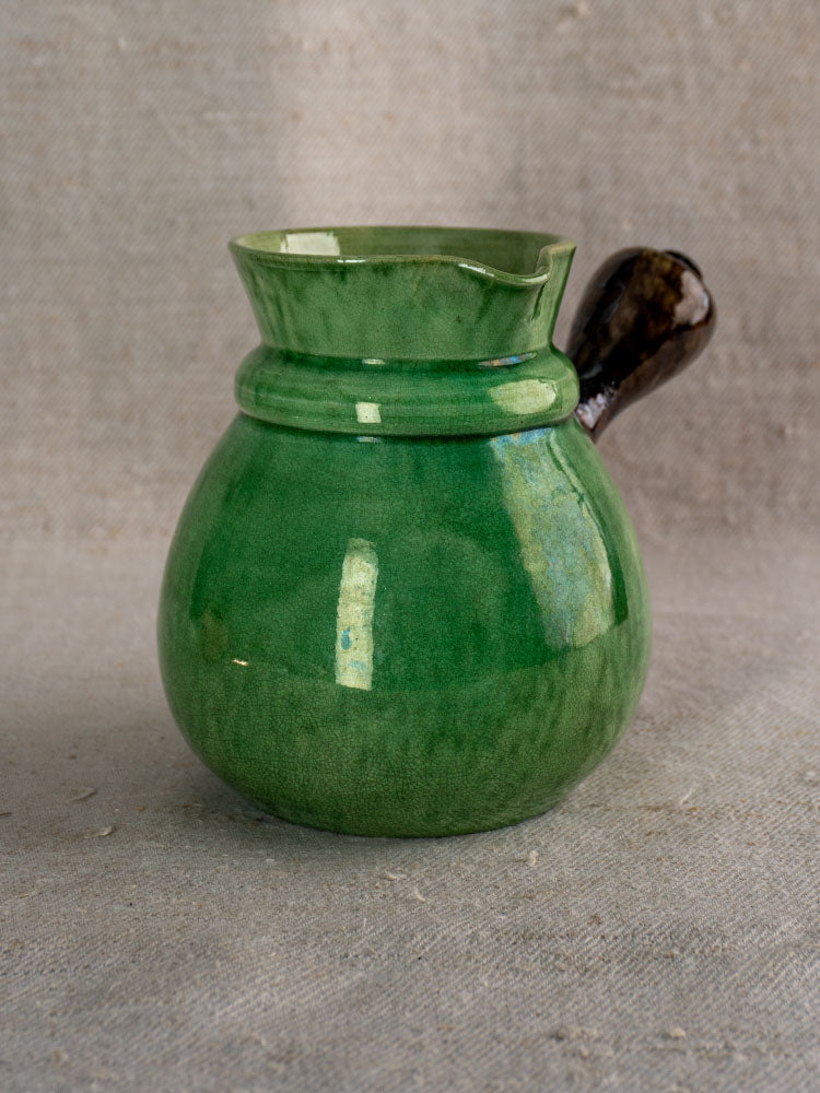 Antique French Dieulefit green glazed earthenware pitcher with wide lip and dark handle on beige fabric.