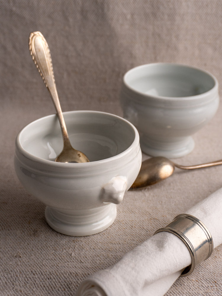Pair of white Antique French Lion Head Soup Bowls, one with silver spoon, on neutral cloth with napkin and cutlery nearby.