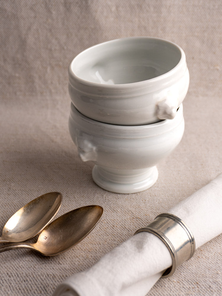 Antique French white porcelain lion head soup bowls, stacked on beige fabric with antique silver spoons and a rolled napkin.