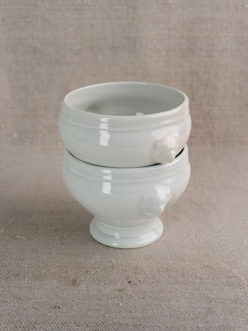 Antique French lion head porcelain soup bowls with decorative handles, stacked on beige textured fabric.