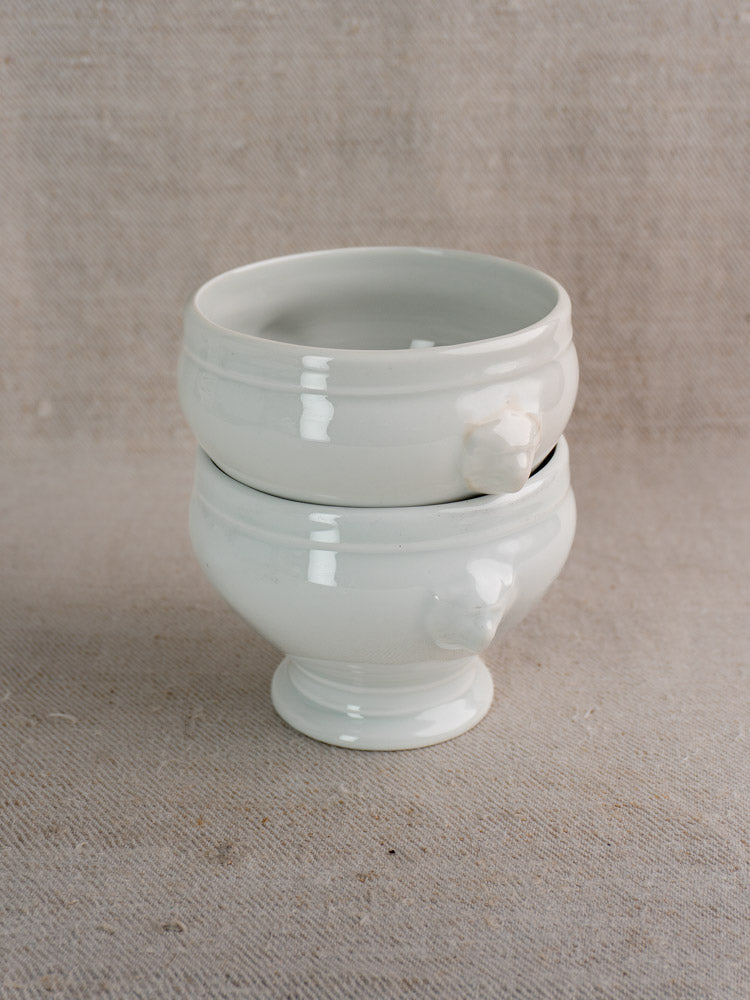 Antique French lion head porcelain soup bowls with decorative handles, stacked on beige textured fabric.