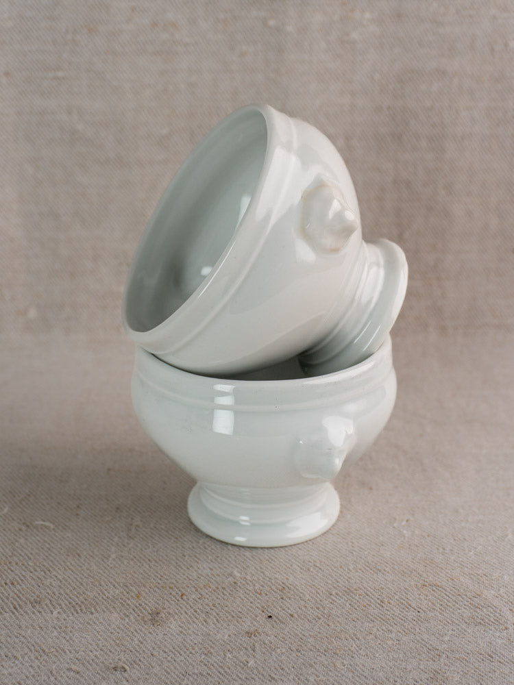 Antique French lion head soup bowls in white porcelain, stacked on textured beige fabric with neutral background.