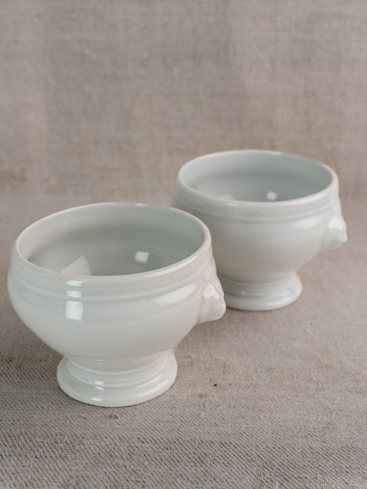Pair of antique French lion head white porcelain soup bowls with glossy finish and refined handle detailing on beige textured fabric.