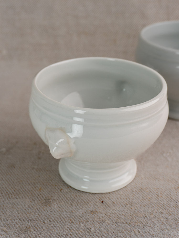 Pair of antique French white porcelain lion head soup bowls with handle, early to mid 20th century, on beige textured surface.