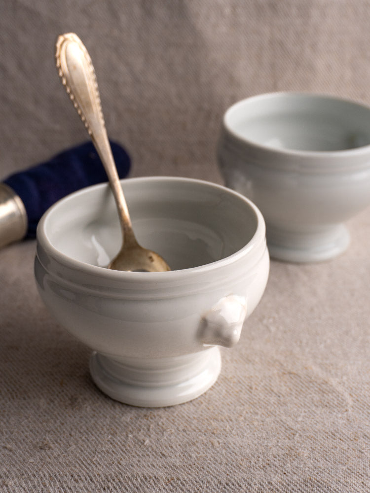 Antique French lion head soup bowls in white porcelain with gold spoon, displayed on a beige surface with blue napkin and silver ring.